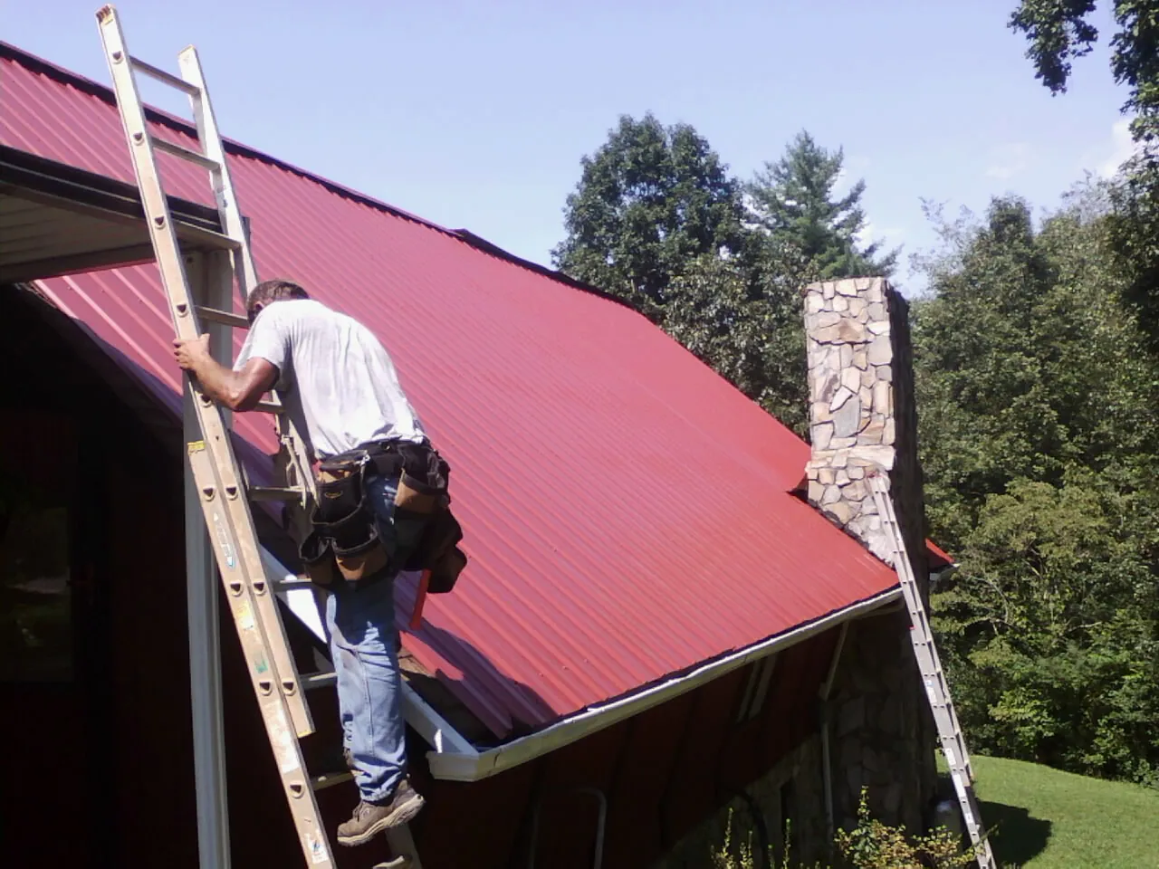 Roofer on a ladder beside a red metal roof and stone chimney — Soffit & Fascia Repair by Roofing Brothers in Yankeetown